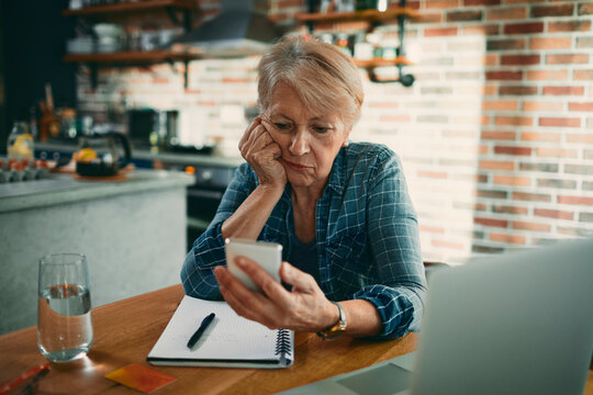 Senior woman looking at smartphone with bills in home kitchen