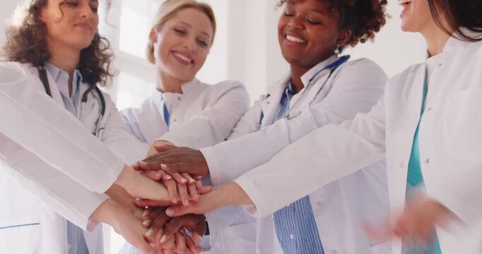 Team of happy diverse female doctors stacking their hands and having fun. Group of smiling multiracial women in medical uniforms pile up their hands and applaud each other. Healthcare teamwork concept
