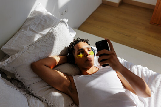 Man relaxing in bed with eye patches and phone