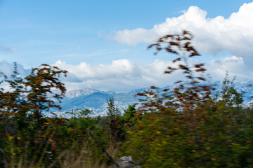Obraz premium Blurred wild plants in foreground with layered karst mountain range and dramatic clouds viewed through car window during road trip in Croatia. Concept of motion, speed and fleeting travel moments.