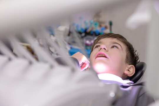 Young boy lying in dental chair with mouth open, looking up at dental light and equipment in modern clinic setting