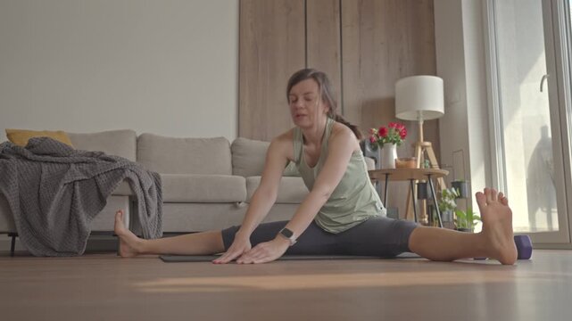 Woman in sportswear sits in straddle position and stretches forward on mat. Concept of flexibility training and healthy lifestyle