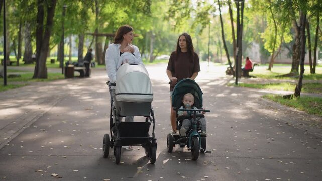 Park stroll lifestyle two mothers walking along sunlit treelined path pushing strollers and tricycle, exchanging conversation and laughter, relaxed weekend bonding and parenting moment in warm natural