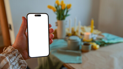Hand holding a smartphone with a blank white screen in front of a blurred table setting