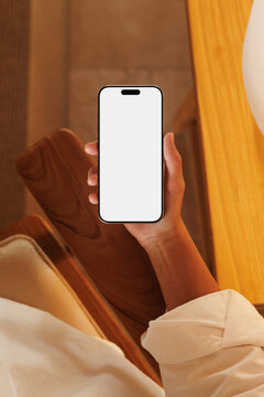 Hand holding smartphone with blank mockup screen on chair 