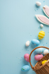 Fototapeta premium Happy Easter vertical scene featuring a pastel painted egg in a wicker basket with bunny ear cookie decorations and scattered confetti on a soft blue background evoking spring celebration