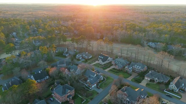 American suburban neighborhood featuring traditional brick houses, manicured lawns, and quiet streets during a golden hour sunset. Aerial cinematic wide-angle video.