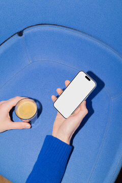 Hand holding smartphone with white screen and coffee cup on blue chair