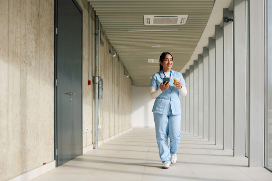 Happy nurse using smart phone while walking with cup of coffee through hospital hallway.