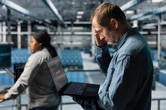 Data center software developer upset after receiving critical error popup on laptop display. Frustrated man in server hub alerted by bugs causing low hardware performance