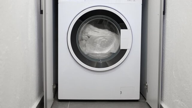 Modern washing machine spinning clean laundry. White clothes and laundry spinning inside the drum of a modern washing machine during a wash cycle. Household appliance working to clean textiles