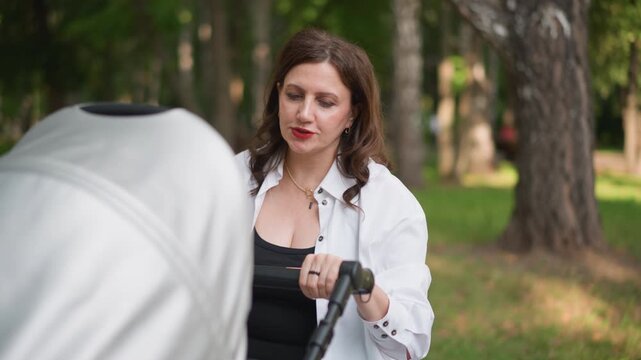 Caucasian mother checks stroller handle in park, thoughtful expression, glance at wrist, planning walk schedule, focus on safety and protection, routine decree shaping daily lifestyle