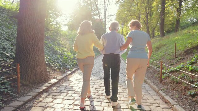 Seniors caucasian walking sunlit cobblestone path supportive trio arm in arm casual clothes dappled light through trees relaxed pace friendship outdoor leisure summer afternoon