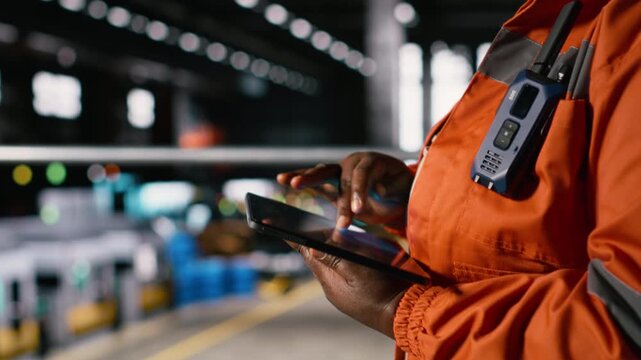 Black female worker evaluates heavy equipment activity on tablet, in a busy engineering plant. Machinery hall filled with steel work, welding activity and mechanical systems for heavy industry.