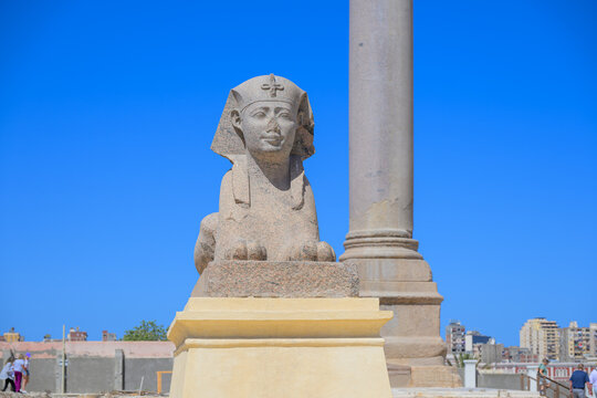 Front view of the pink granite Sphinx with Pompey's Pillar behind at the ancient Serapeum Complex in Alexandria, Egypt.