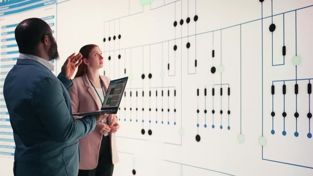 Team of technicians review PCB layout, using laptop to project it on large digital screen. Teamworking RD startup colleagues analyze electrical circuit connections for electronic device manufacturing