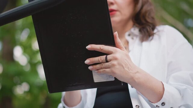 Caucasian woman holding decree document closeup of hands and rings, black folio on clipboard, presenting mandate to audience, formal tone, outdoor garden backdrop, authoritative lifestyle moment