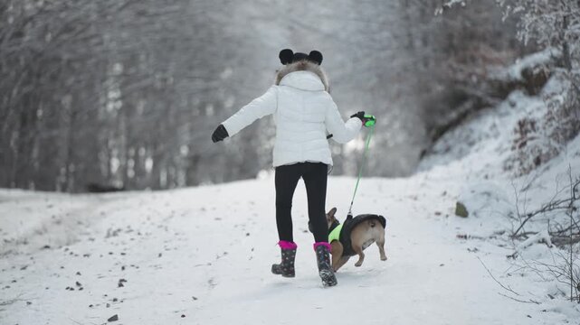 Young teen with funny hat and boots running with french bulldog dog in the mountain forest winter snow landscape, slow motion