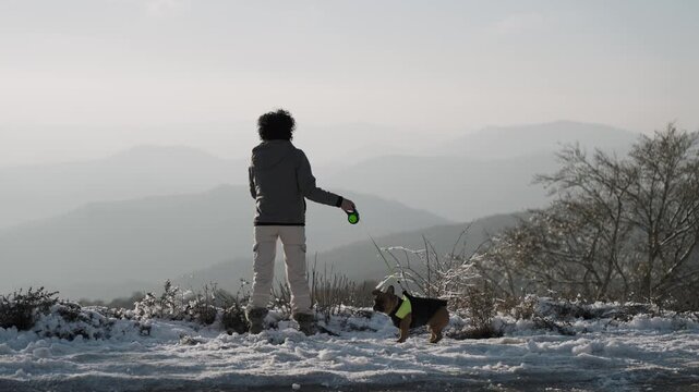 Curly hair woman with french bulldog dog on leash standing on winter snow covered view point enjoyng mountain landscape scenery at sunset