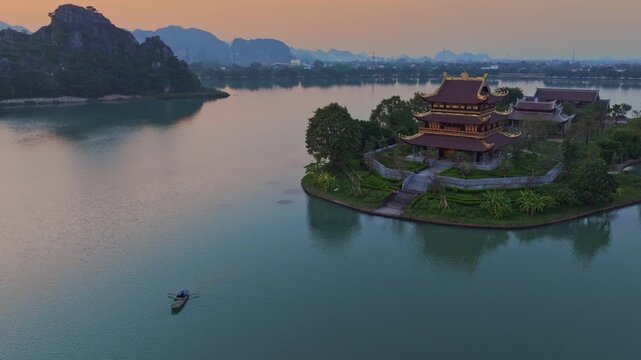 A lone rower navigates the tranquil waters of a lake, Ninh Binh, Vietnam, at dawn, with the majestic Chua Vang Pagoda standing proudly on its island.