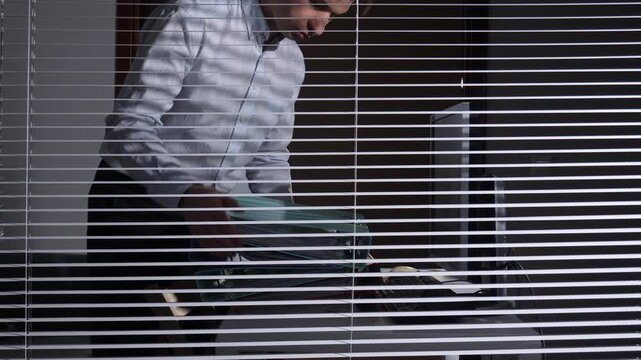 Businessperson carrying a stack of files and folders into a private office, seen through the slats of venetian blinds, symbolizing workload, confidentiality, and focused work