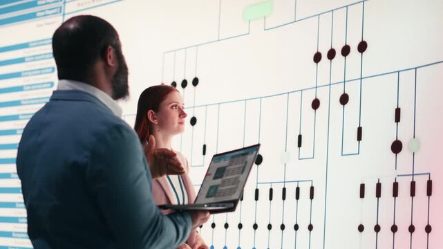Team of technicians review PCB layout, using laptop to project it on large digital screen. Teamworking RD startup colleagues analyze electrical circuit connections for electronic device manufacturing