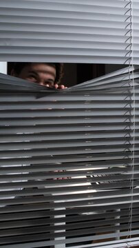 Vertical video person peering through venetian blinds into a dim room, eyes and shadowed face visiblesuggesting surveillance, secrecy, privacy invasion, or cautious curiosity