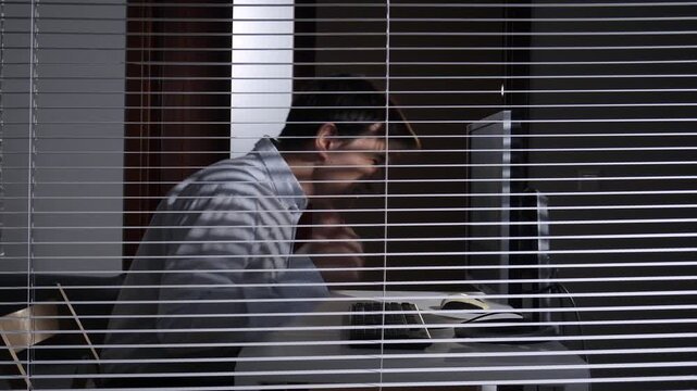 Overworked businessman feeling frustrated and angry while working hard on a desktop computer in an office, seen through window blinds at night, depicting stress, pressure, and overwork