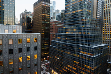 A high-angle view of Manhattan, New York City, showcasing a dense cluster of modern skyscrapers lining a busy street below. Tall glass and steel office towers rise on both sides, their illuminated win