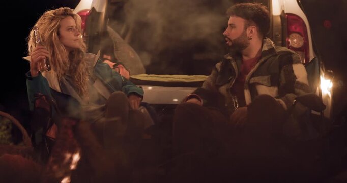 Two friends share stories by the campfire while enjoying a night outdoors in a remote location with a car in the background and a glowing fire pit