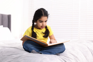 Fototapeta premium Cute little girl reading book on bed at home