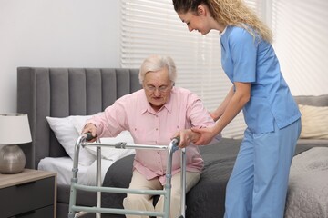 Fototapeta premium Care worker helping senior woman with walker to stand up from bed indoors