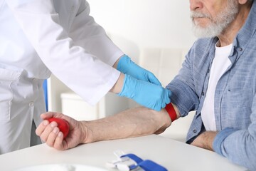 Doctor putting medical tourniquet on senior man's arm in clinic, closeup