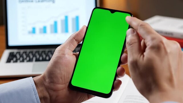 A person's hands holding a smartphone with a green screen, with a laptop displaying a business chart in the background.