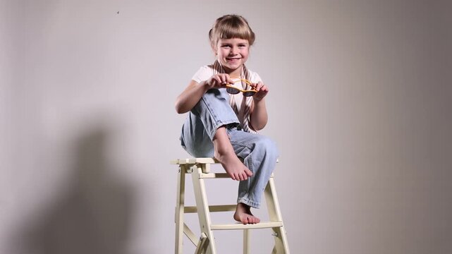 Fashion. Cute little girl in stylish outfit on ladder against light background
