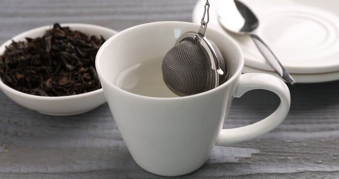Brewing tea. Woman putting metal infuser with dry leaves into cup with water at grey wooden table, closeup