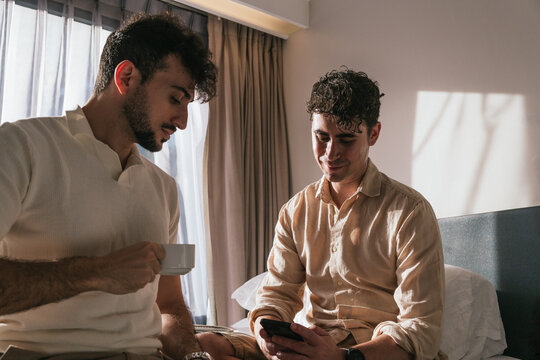 Young men relaxing in bed during morning, one drinking coffee and other using smartphone, sharing a moment of togetherness