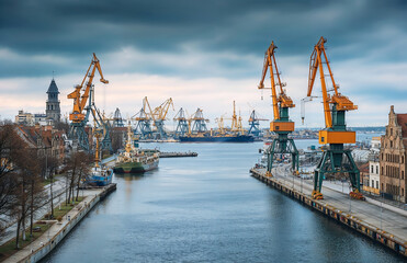 Obraz premium Industrial port with large cranes and boats on river under dramatic cloudy sky