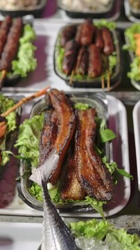 Vertical screen Grilled Fish Pork Belly Street Market Closeup Of Glazed Smoked Pork Belly Slices On Bed Of Lettuce With Skewered Sausages And Mackerel Tail On Ice, Vendor Display, Charred