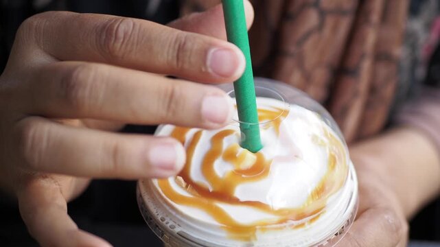 Close up shot of person hands holding frappe coffee with caramel drizzle, slowly pushing straw through lid into drink on sunny day