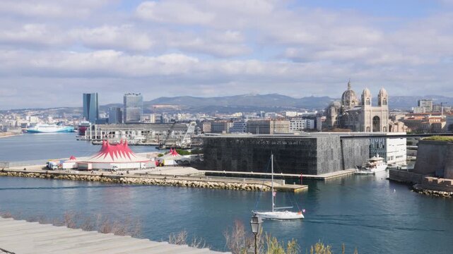 Wide view of Marseille harbor entrance with MuCEM and Cath&eacute;drale La Major in frame. A sailboat exits the port, city visible in the background under a cloudy sunny sky.