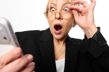 Surprised middle aged woman in glasses looks shocked at smartphone screen. Isolated on white...
