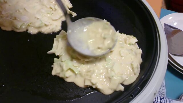 Cooking Japanese Okonomiyaki Savory Pancake on a Hot Griddle. Chef preparing traditional Osaka-style okonomiyaki with fresh cabbage batter on a flat iron griddle at a street food market.