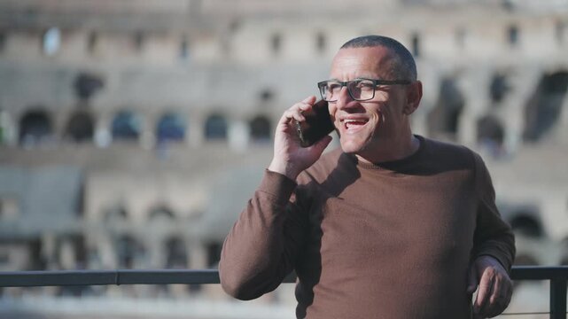 Portrait of young businessman making a phone call smiling at sightseeing visit of Colosseum in Rome. Slow motion cinematic shot with selective focus