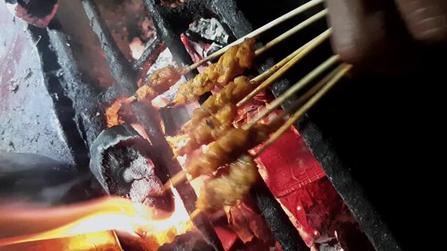 Indonesian Satay Grilled over Charcoal Fire. Traditional Indonesian street food, satay skewers being grilled over hot charcoal fire with smoke at a local night market.