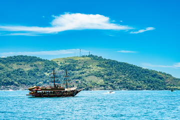 Obraz premium ketch-rigged wooden tourist boat sailing along Angra dos Reis coastline, Brazil. Features rainforest-covered mountains and traditional sailing charm for travel and documentary projects.