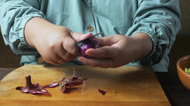 A woman's hands peel a red onion with a knife on a cutting board