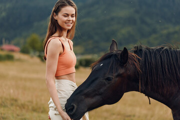 Obraz premium A smiling young woman stands in a sunlit field beside a dark horse, wearing an orange crop top and light shorts. The outdoor moment radiates calm, friendship, and natural beauty.