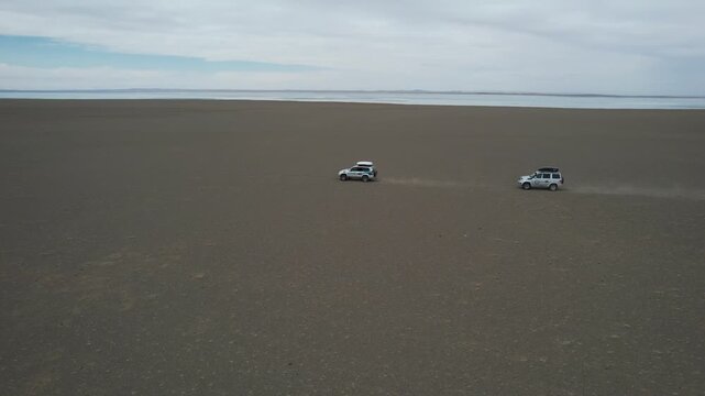 Two White Suvs Traverse Barren Salt Flat Under Overcast Sky, Leaving Low Dust Trails While Remote Expedition Crosses Endless Plain, Muted Horizon And Wide Open Space Convey Solitude And Endurance
