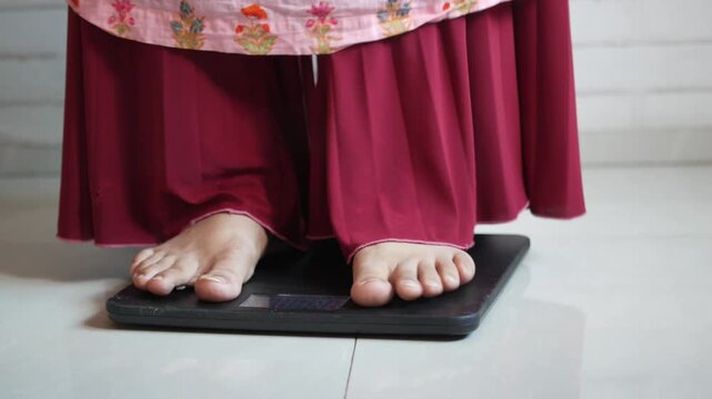 Woman stepping on weighing scale to check body weight at home, close-up on feet